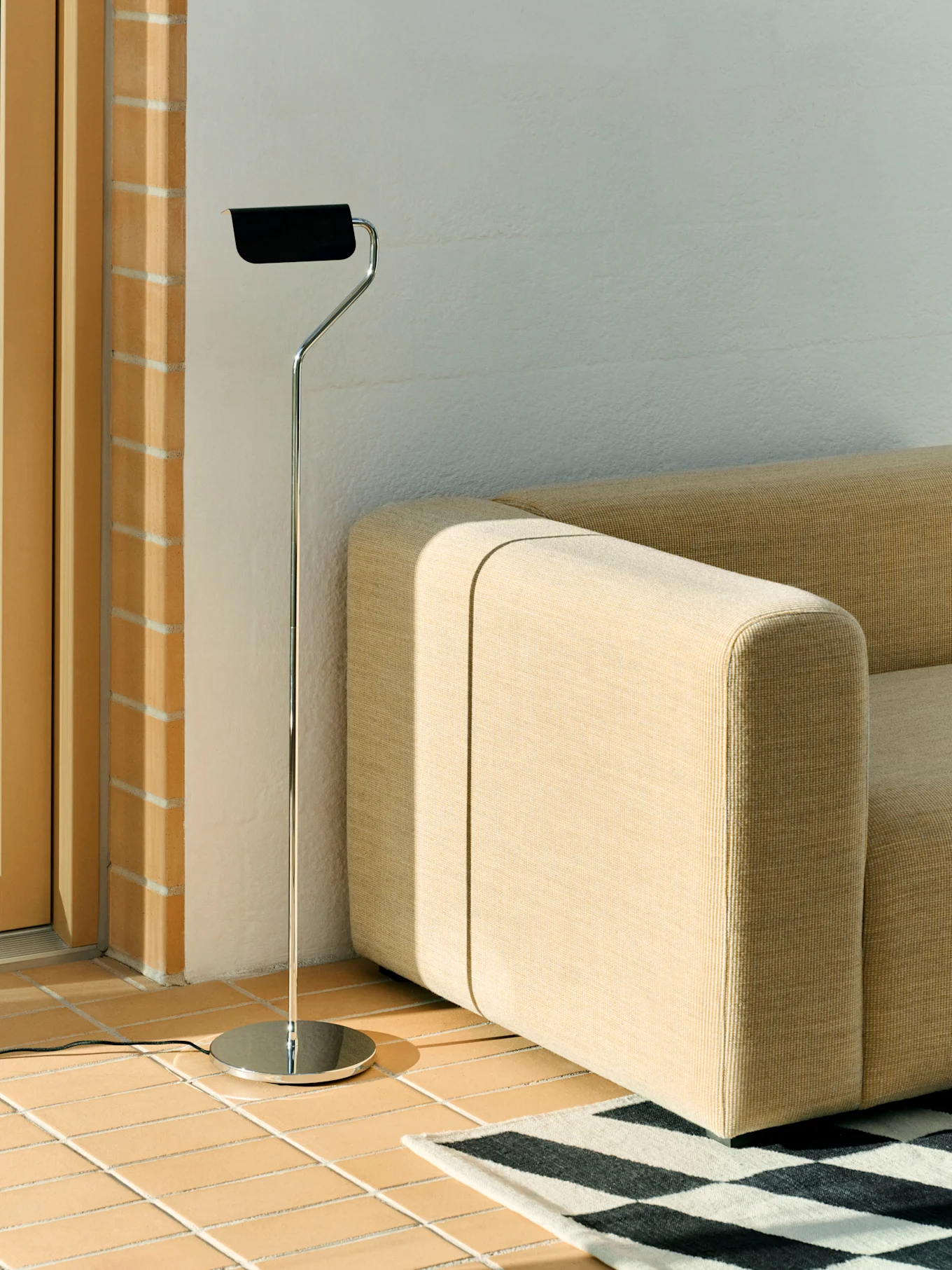 A chrome floor lamp with a black shade stands next to a beige modular sofa and a black and white geometric rug on a tiled floor.