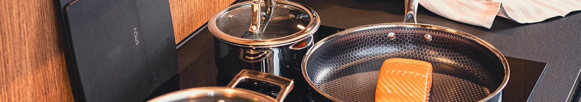 Stovetop view: Salmon in a frying pan, next to a pot. Cookware has a hexagonal pattern and copper trim. 'hâws' logo.