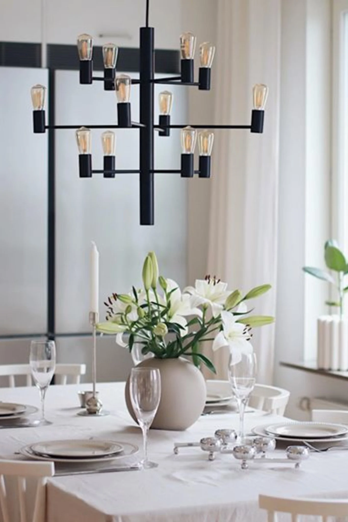 A modern dining room with a table set with white plates, clear glasses, a vase of white lilies, and a tall candle, all beneath a multi-tiered black chandelier with exposed bulbs.