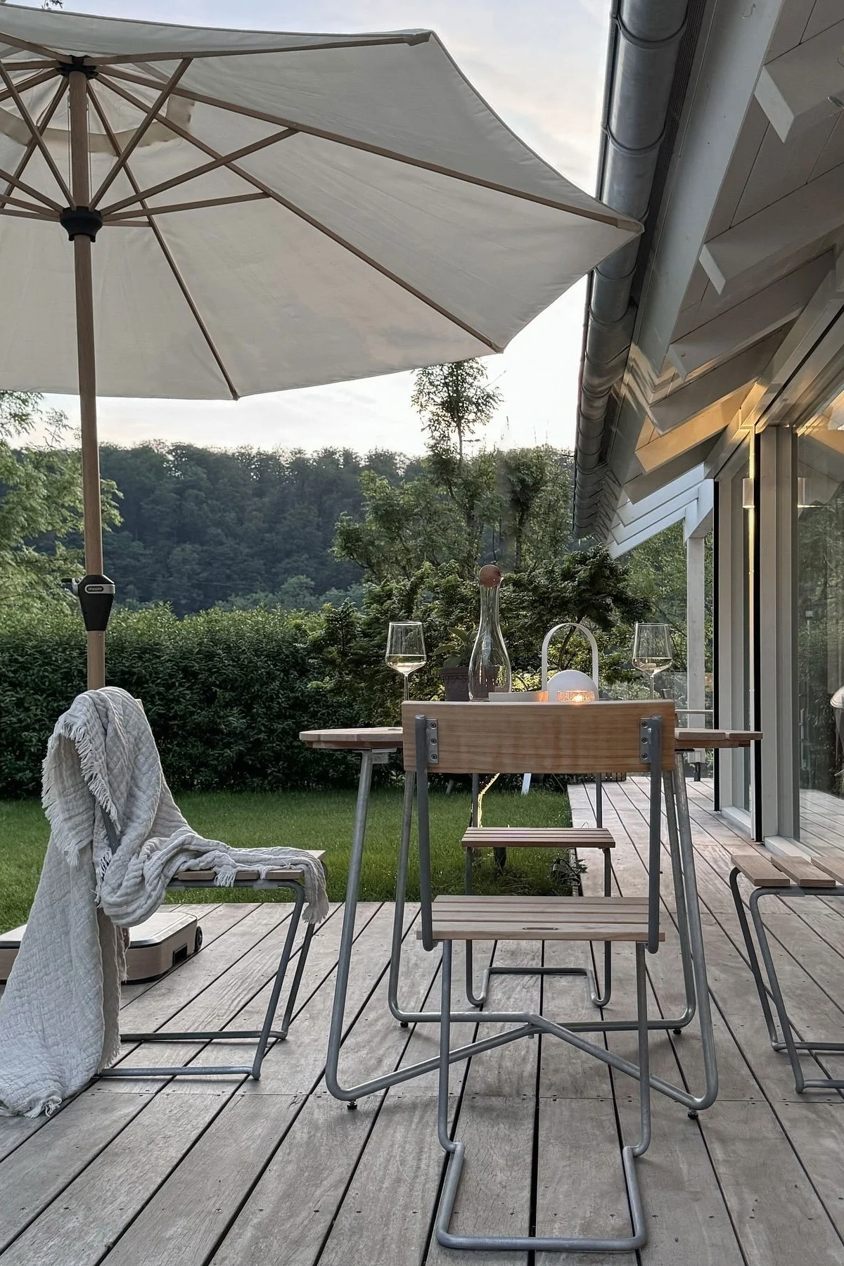 Outdoor patio with a wooden table, two chairs, wine glasses, a carafe, and a lit lantern under a large white umbrella, surrounded by greenery.