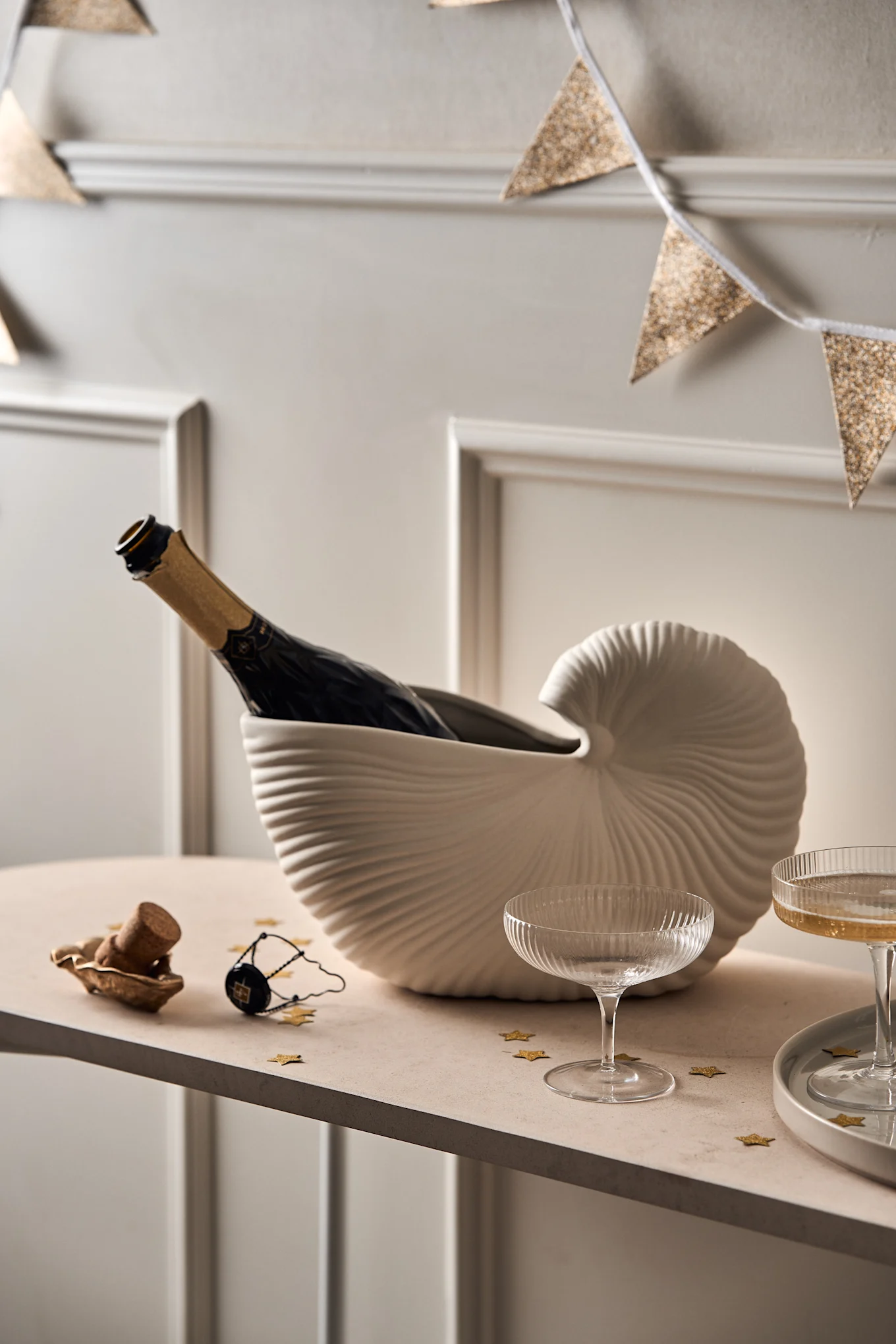 Champagne bottle in a shell cooler, two glasses, cork, and gold star confetti on a table, with a glittery garland.