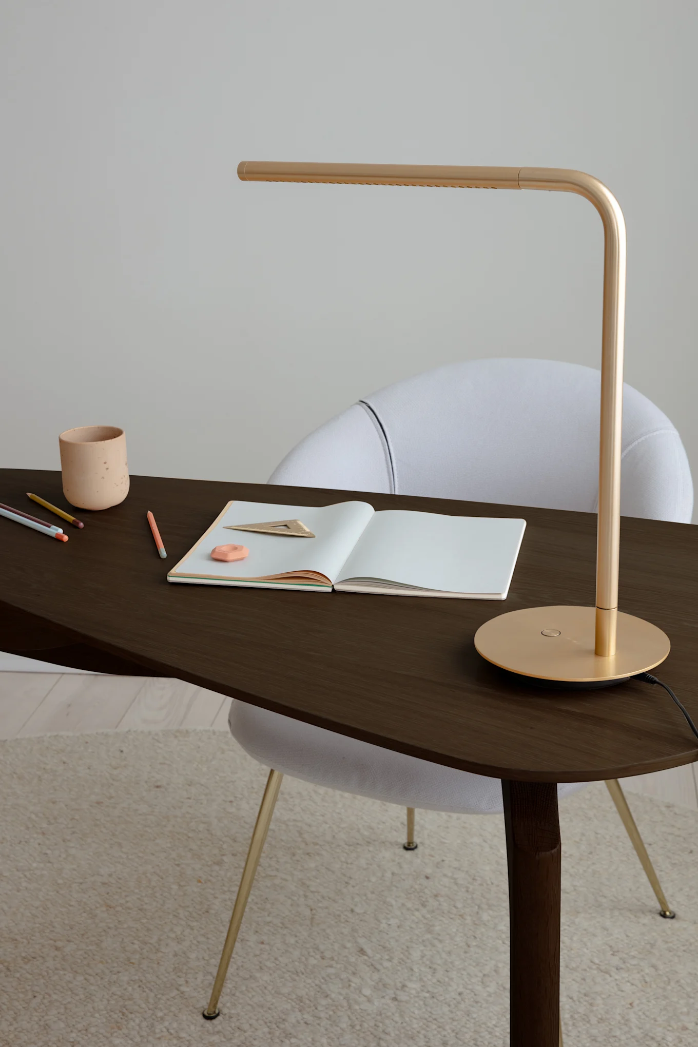 Gold desk lamp on a dark wood desk with an open notebook, pencils, eraser, ruler, and ceramic cup. White chair behind.