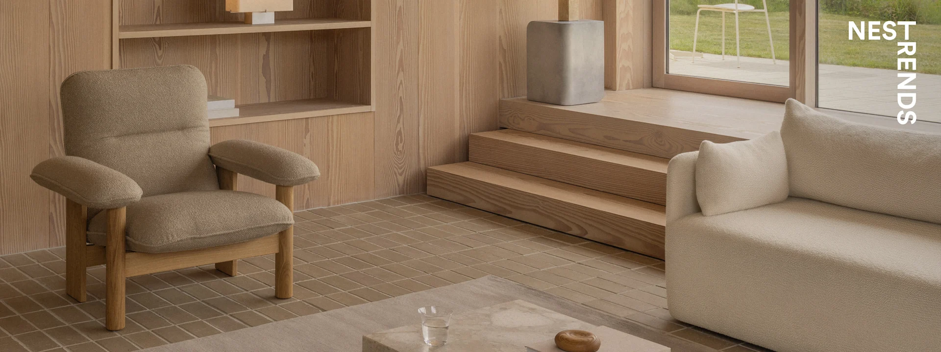 A modern living room with a textured brown armchair, a beige sofa, wooden wall panels, tiled floor, and a view to a garden.