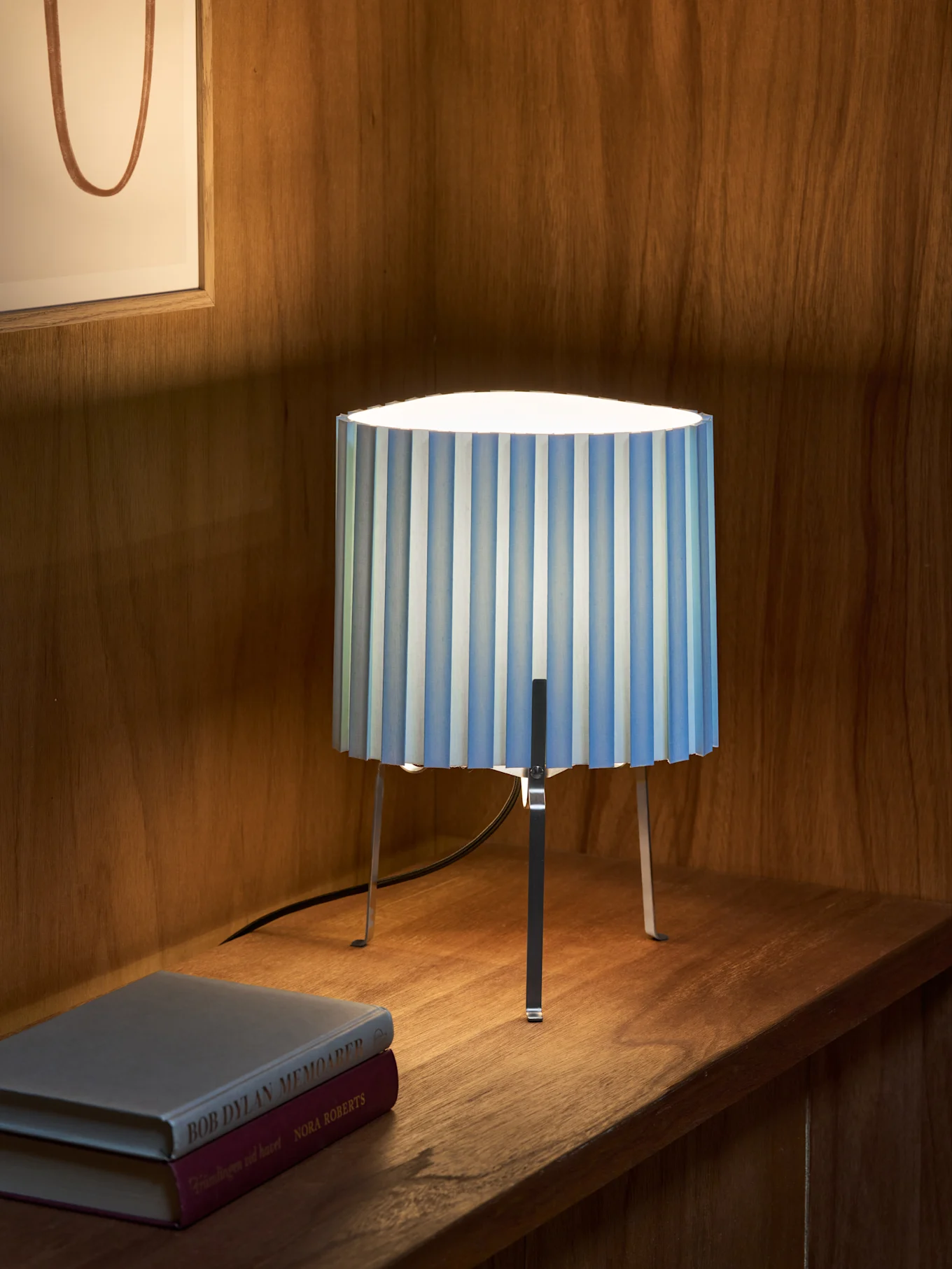 A blue and white striped pleated table lamp with three metallic legs sits on a wooden shelf next to two books. Framed artwork in background.