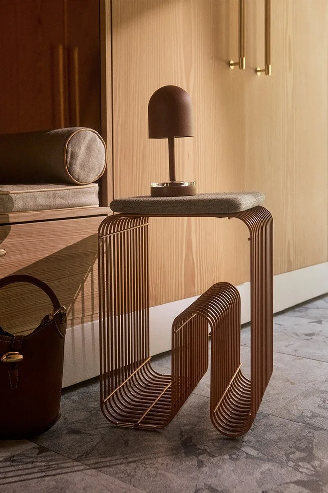 Modern side table made of brown vertical metal rods with a brown dome lamp and handbag, against a wooden cabinet.