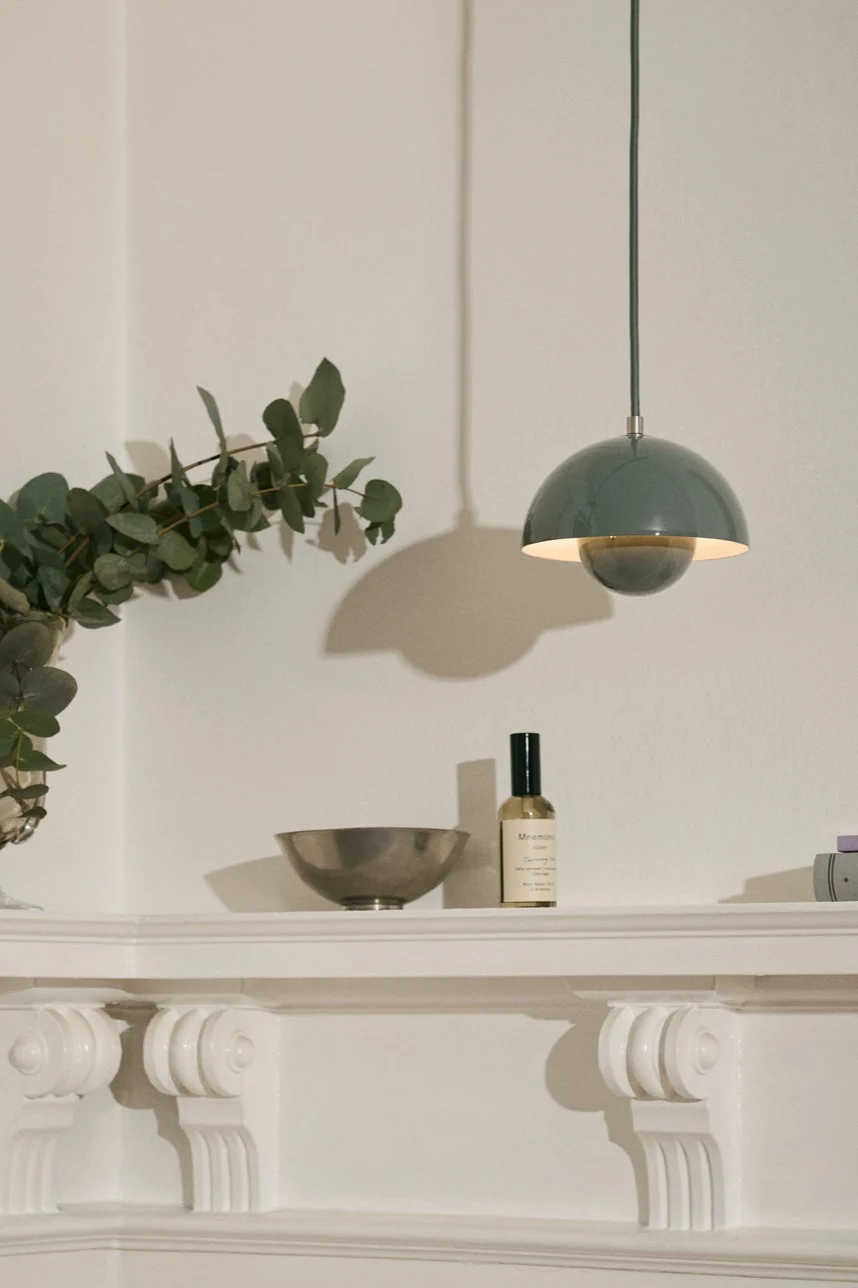 A light green dome-shaped pendant lamp hangs above a white shelf with a silver bowl, a spray bottle, and eucalyptus branches.
