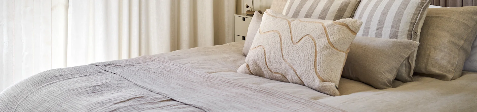 A bed with neutral-toned linen bedding, striped pillows, and a textured white pillow with wavy beige lines.