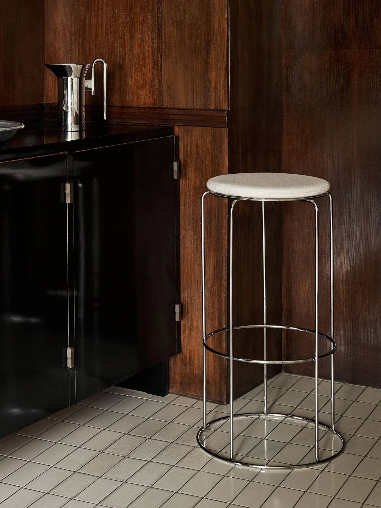 A black cabinet with barware and a chrome bar stool with a light seat, against dark wood paneling and a tiled floor.