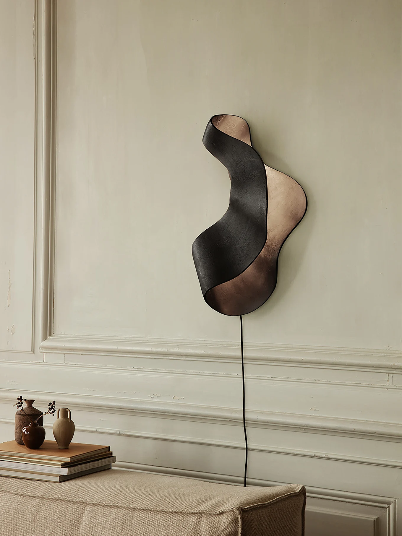 Black and bronze wavy ribbon-shaped wall sconce on a light wall above a sofa with vases and books.