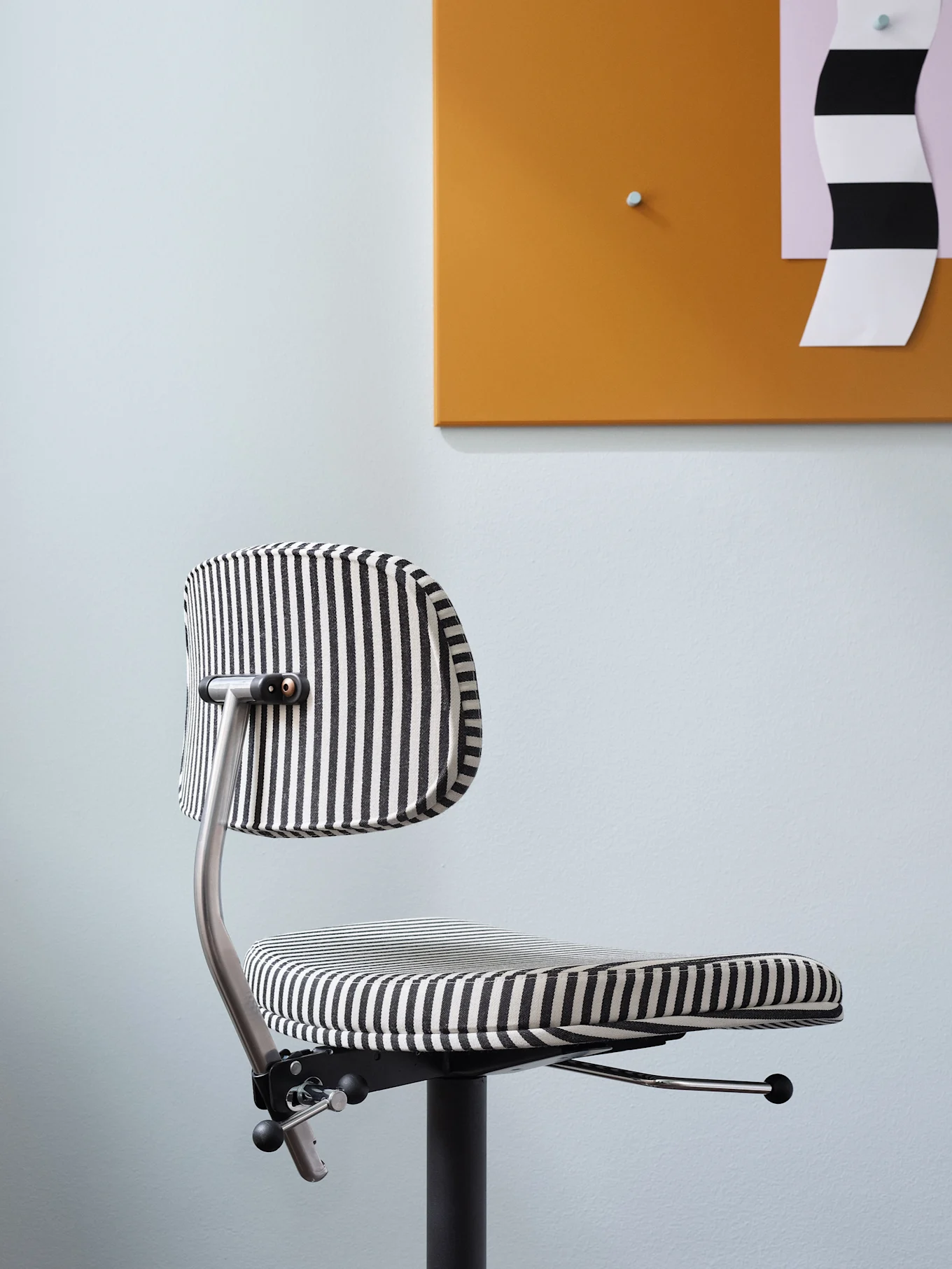 A modern office chair with black and white vertical stripes, set against a light blue wall and an orange board.