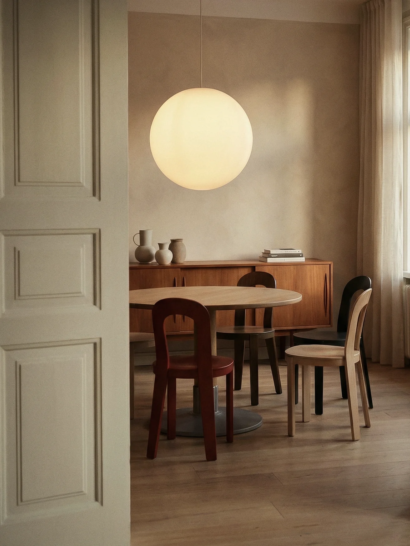 A modern dining room with a round wooden table, assorted chairs, a large spherical pendant light, and a credenza with decorative vases.