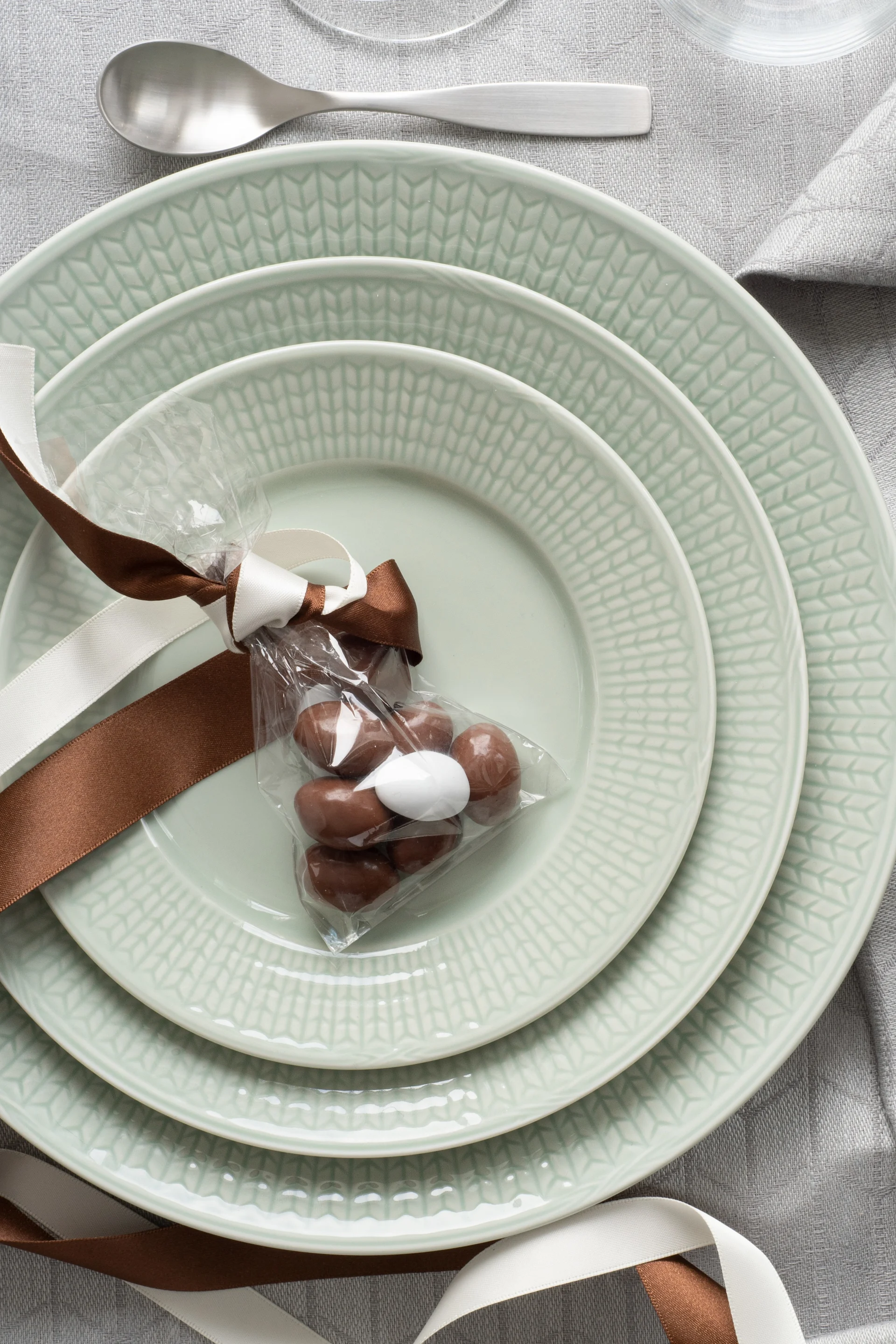 Stacked light green plates with a gift bag of chocolates on a grey tablecloth, beside silver cutlery.
