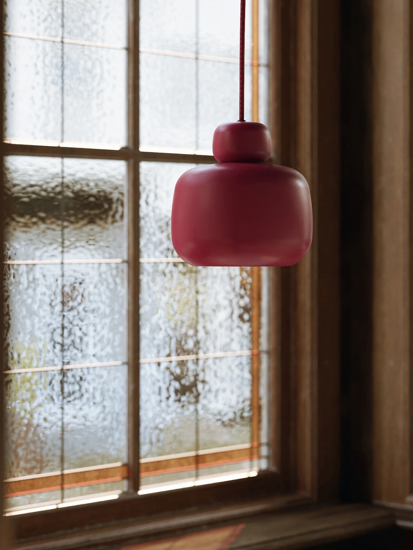 A dark pink pendant light with a two-tiered, rounded shape hangs in front of a textured glass window.