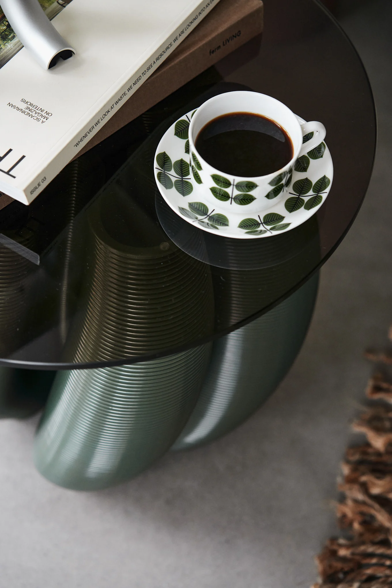 Overhead view of a coffee cup with green leaf pattern on a dark glass table with books and a fluted teal base.