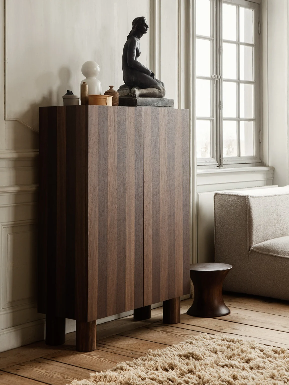 Dark wooden cabinet with vertical stripes, topped by a sitting sculpture, white lamp, and decor, next to a textured sofa and dark stool.
