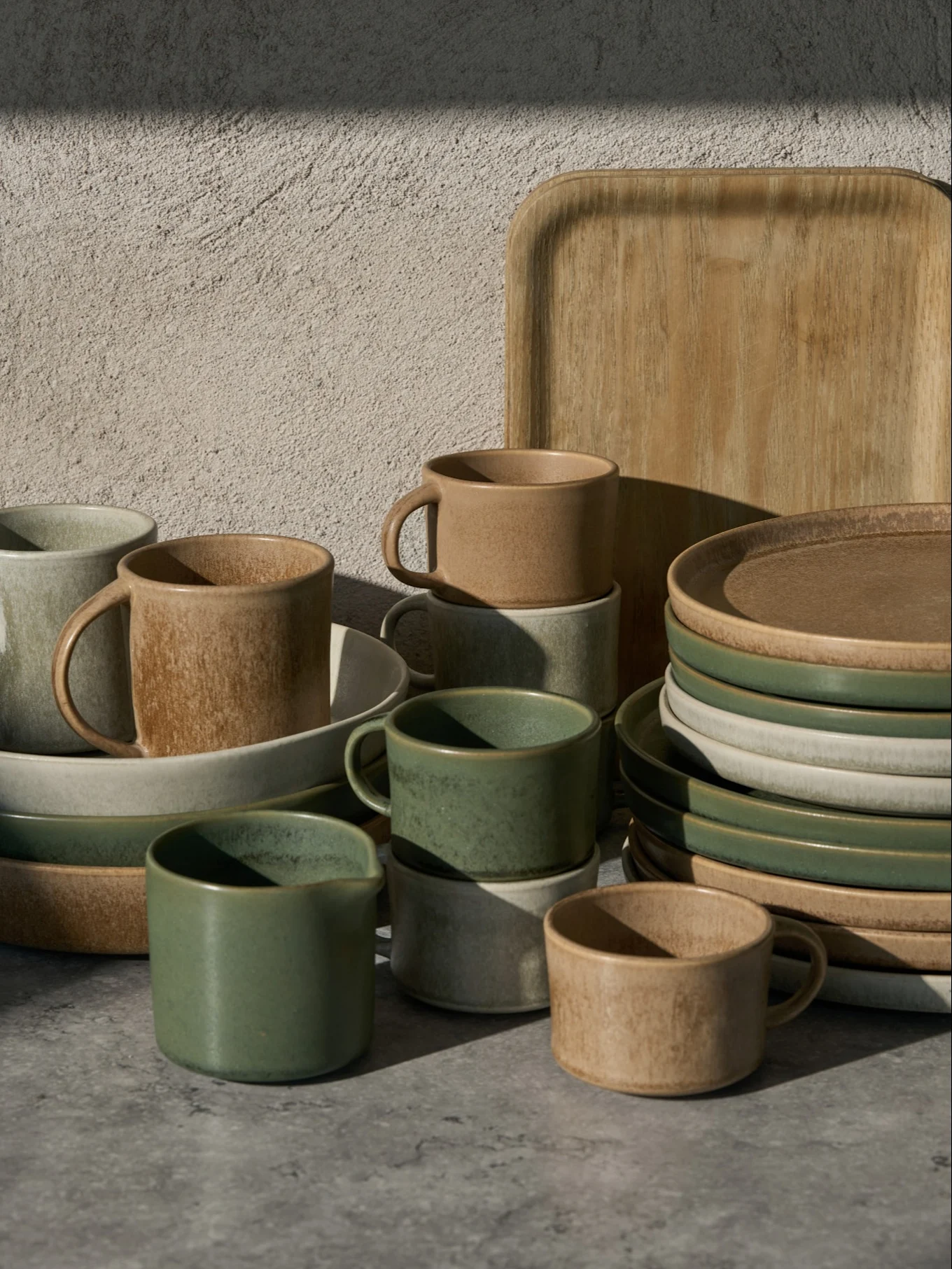 Collection of green, beige, and light gray ceramic cups, bowls, and plates, with a wooden tray, on a gray surface.