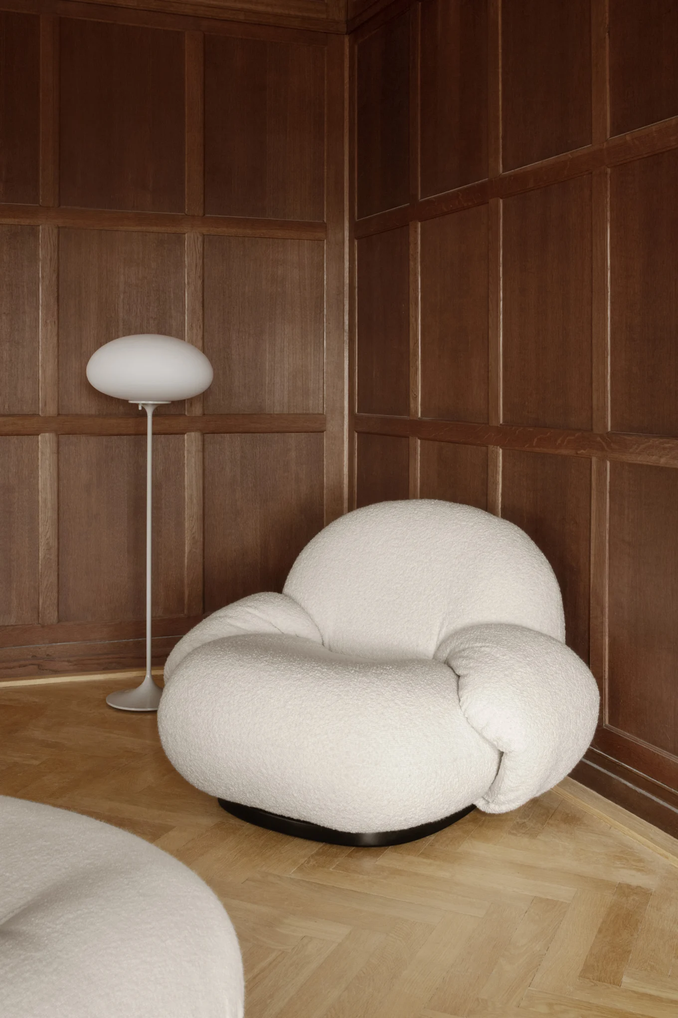 A white boucle armchair and a white oval floor lamp in a room with dark wood paneling and herringbone floor.