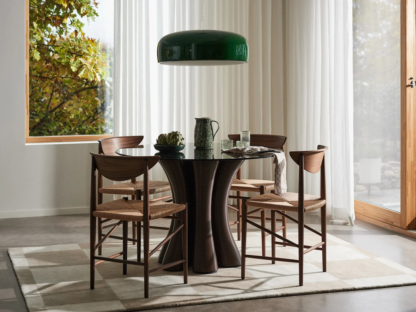 Dining room with dark round table, four wooden chairs with woven seats, green pendant light, and window to a lush garden.