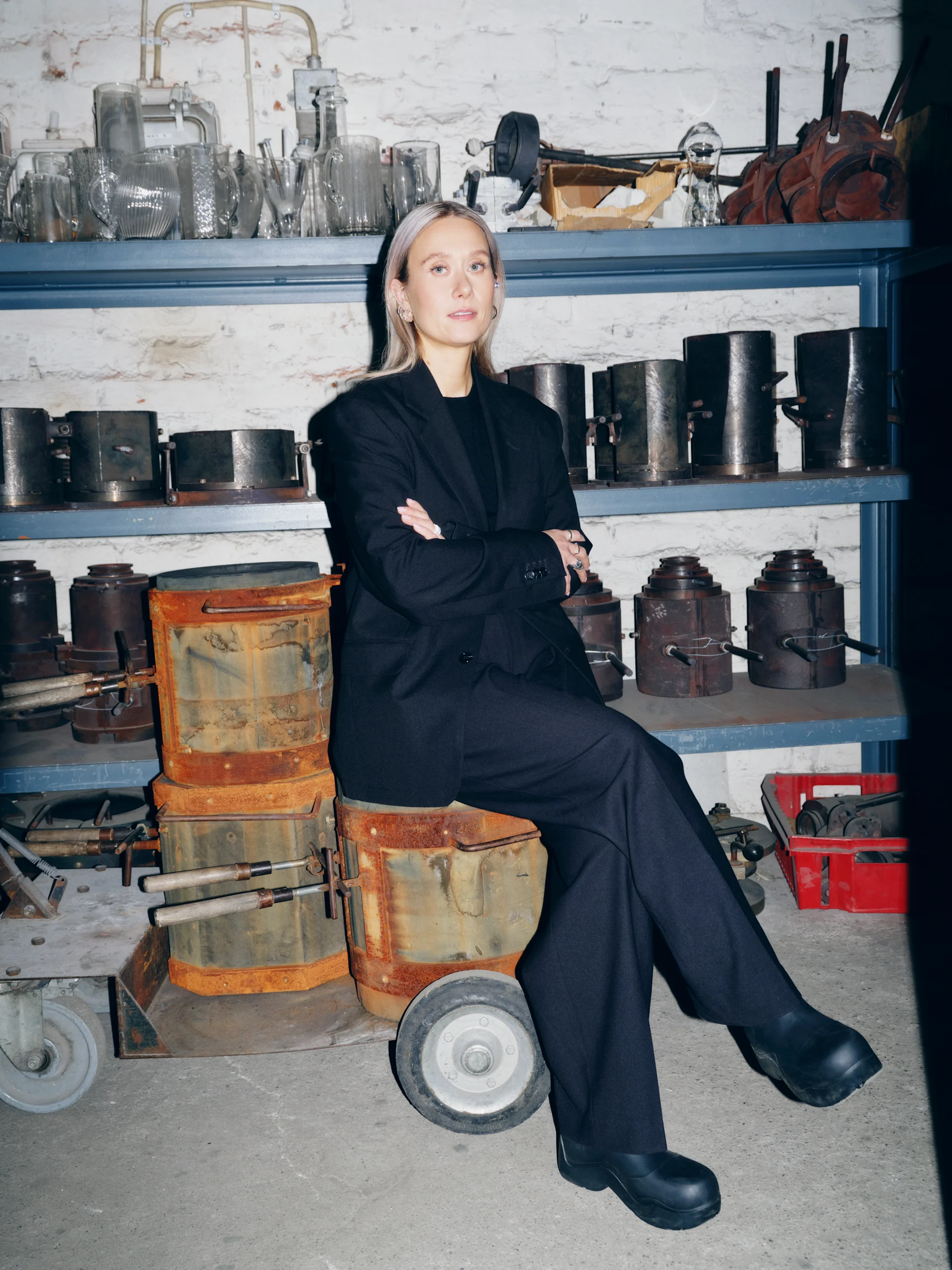 Blonde woman in black blazer and wide-leg pants sitting with crossed arms in an industrial workshop with glass and metal molds.