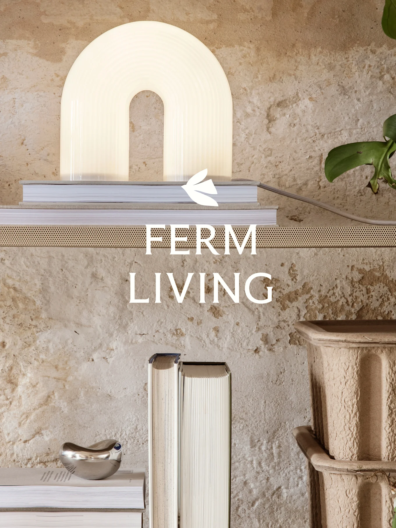 A minimalist interior with a glowing arched lamp, books, and a silver decorative object on a perforated shelf against a textured wall. Ferm Living logo.