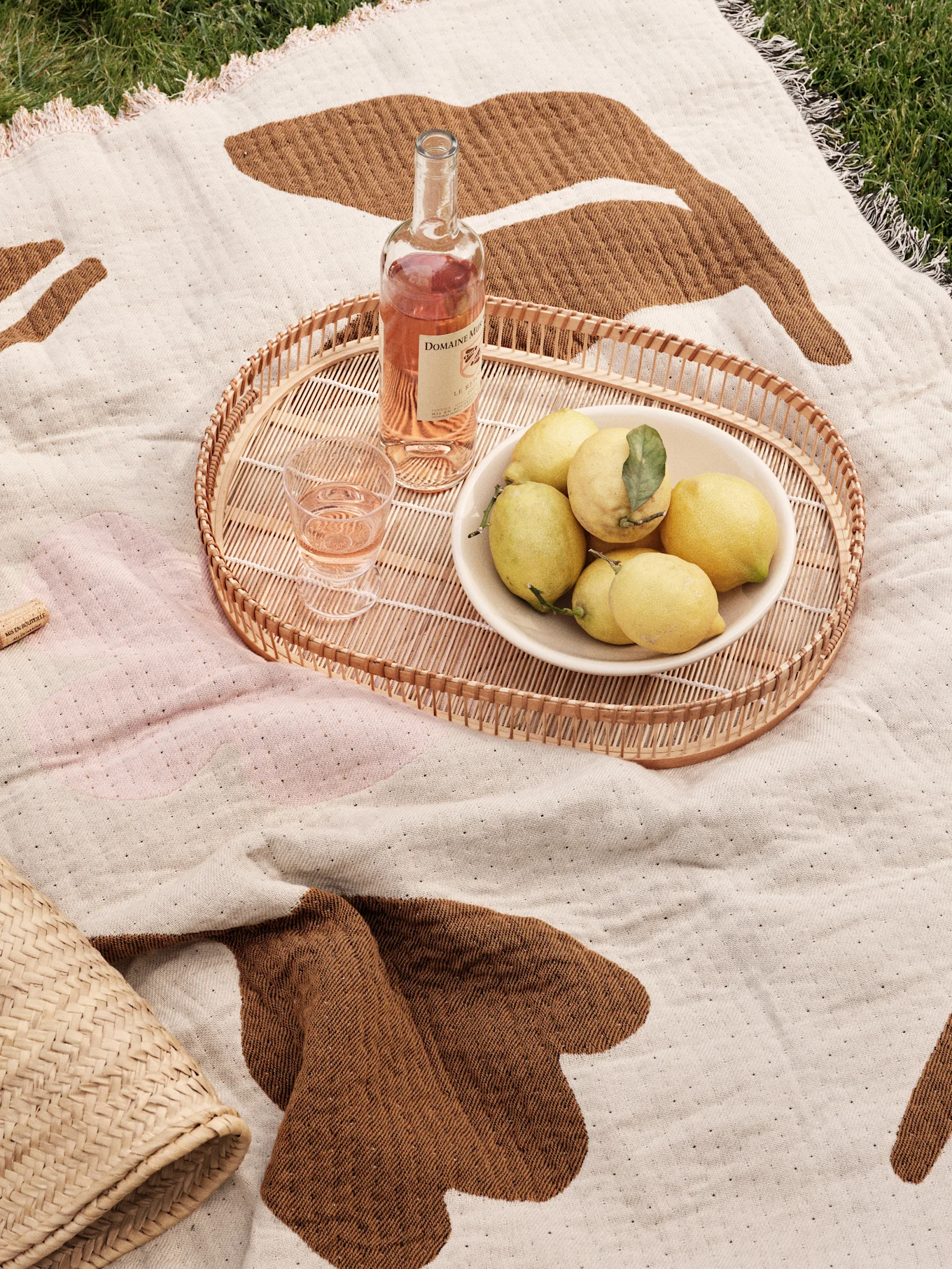 Picnic blanket on grass with rattan tray, rosé wine, glass, and a bowl of lemons.