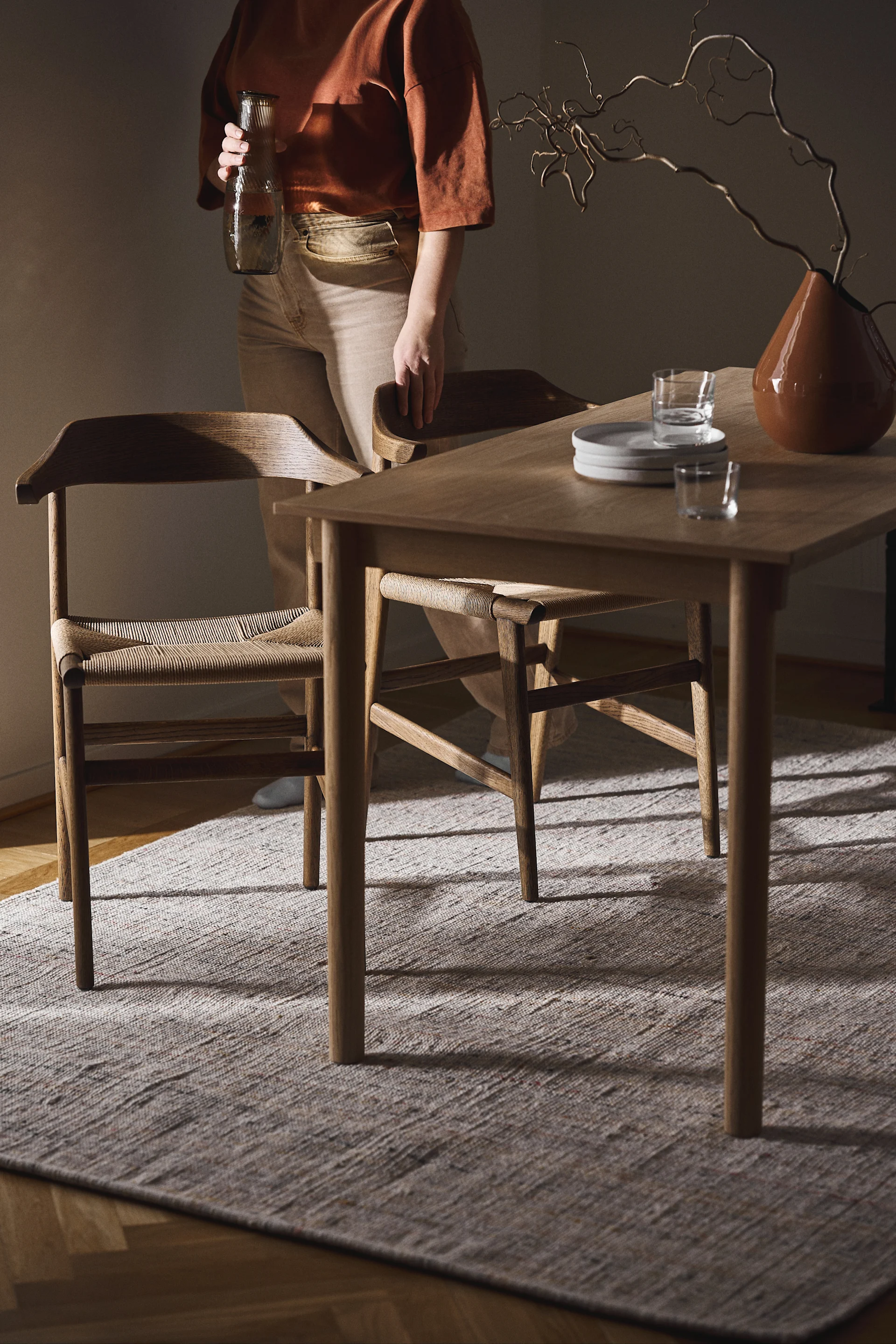 Person holds a ribbed glass carafe next to a wooden dining table with woven chairs, plates, glasses, and a vase of curly branches.