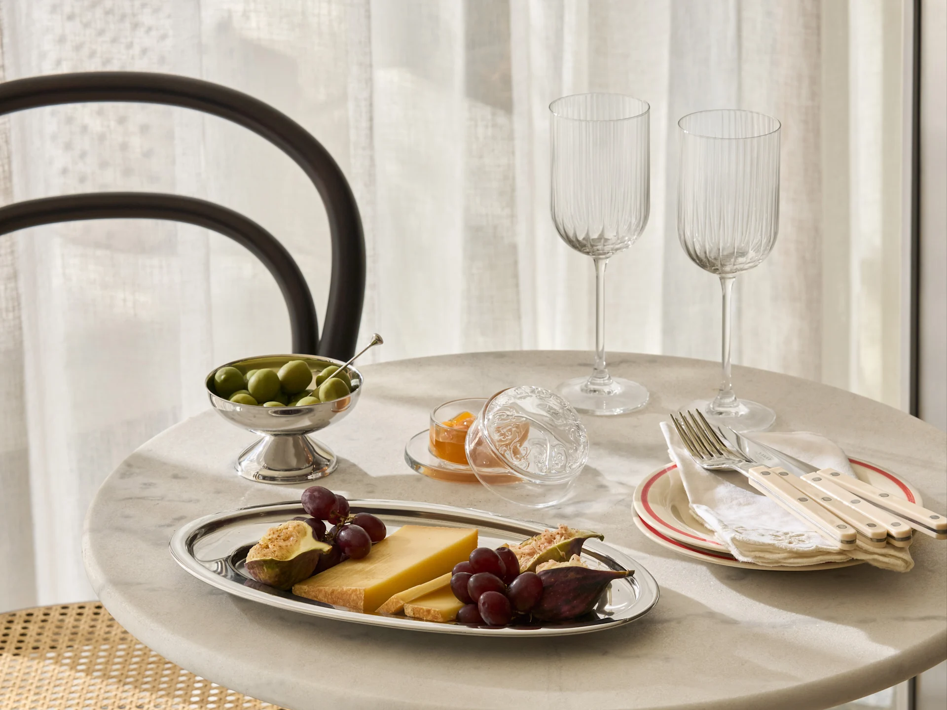 A marble table set with a silver platter of cheese, grapes, figs, a bowl of olives, jam, two ribbed glasses, and cutlery on plates.