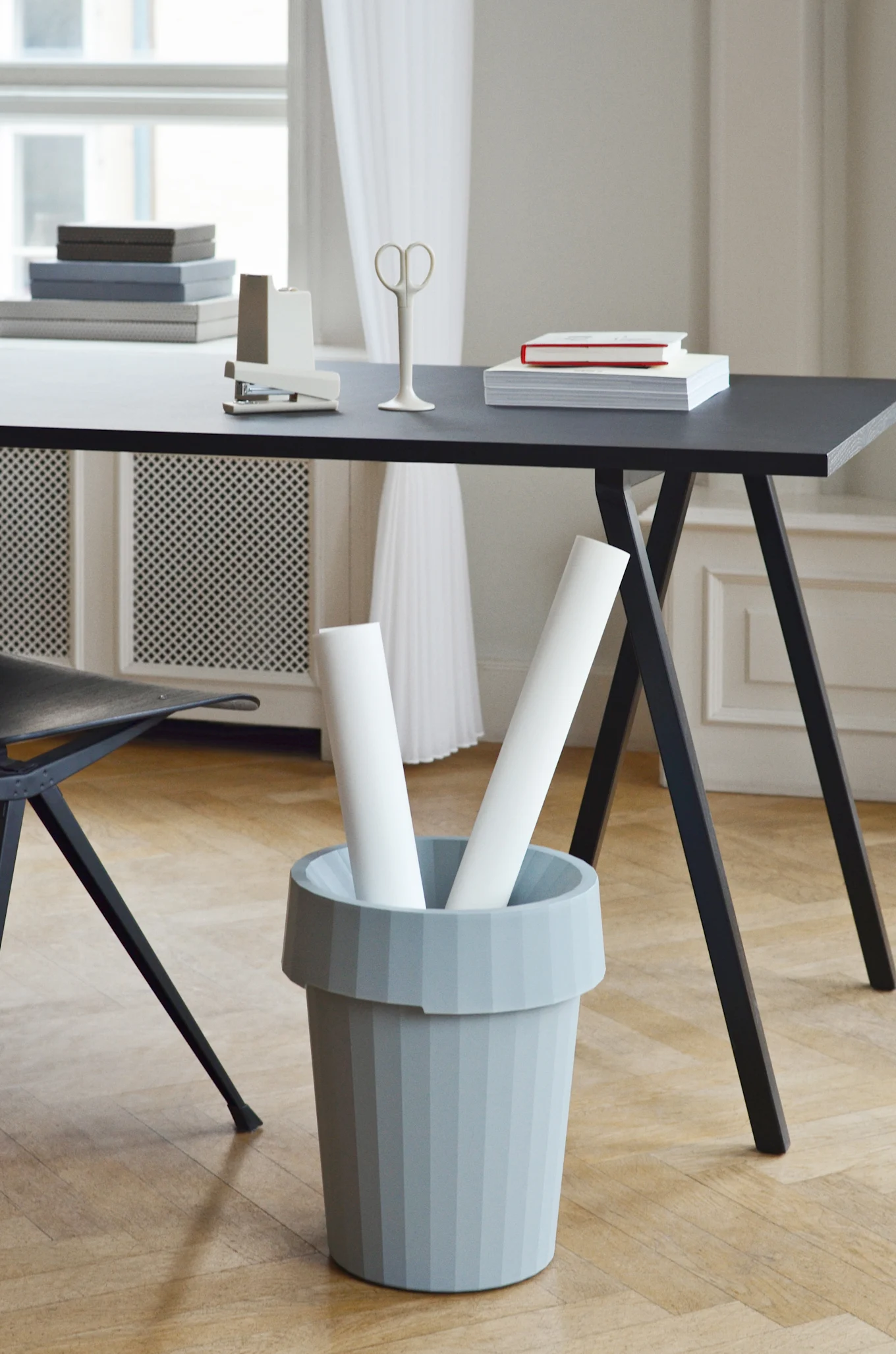 A grey ribbed bin holds two white rolled papers next to a black desk with office supplies in a brightly lit room.