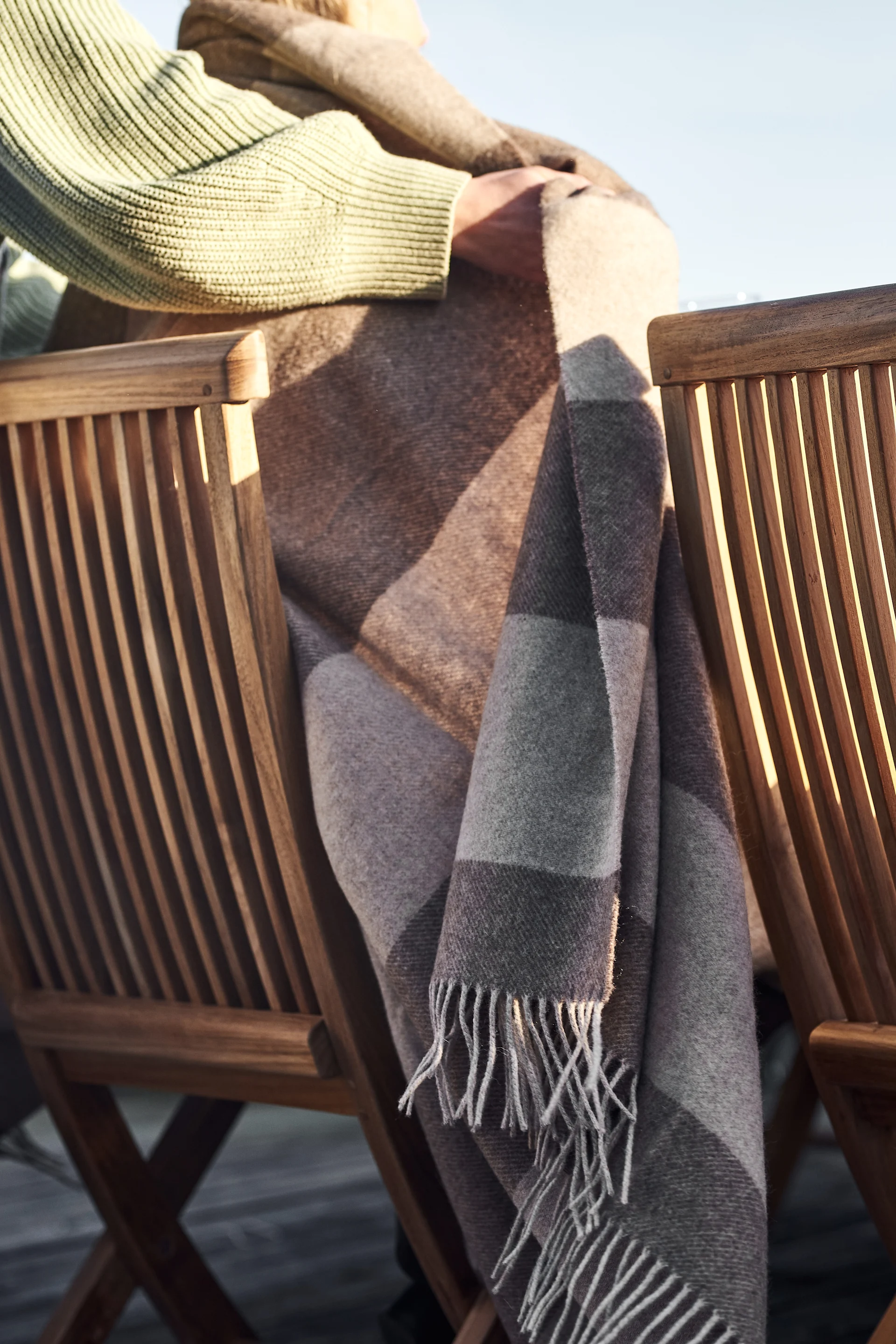 Person in a green sweater wrapped in a brown and gray plaid blanket, sitting in a slatted wooden chair with sunlight casting shadows.