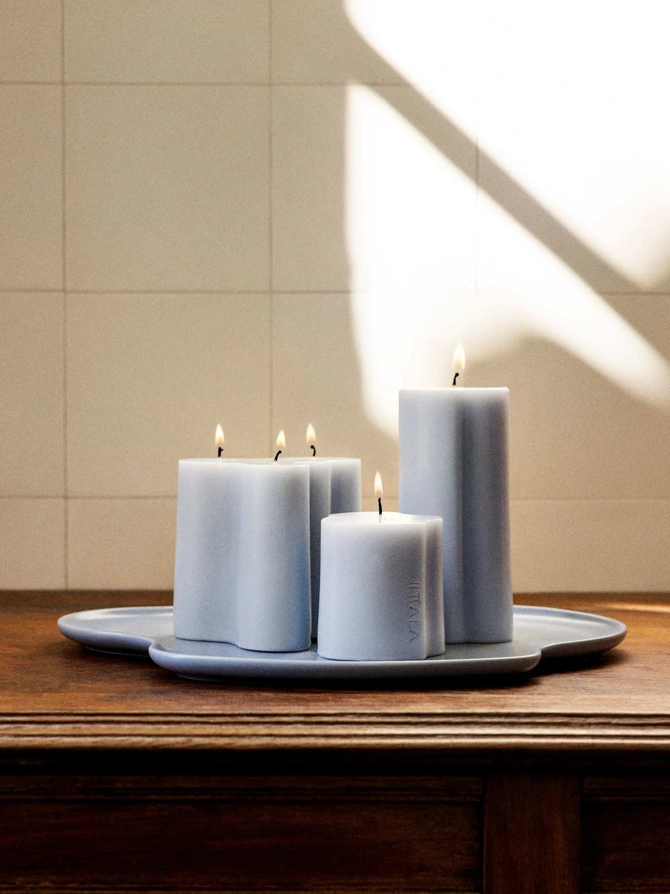 Four lit light blue-grey candles of various shapes and heights on a matching irregularly shaped tray on a dark wooden surface.